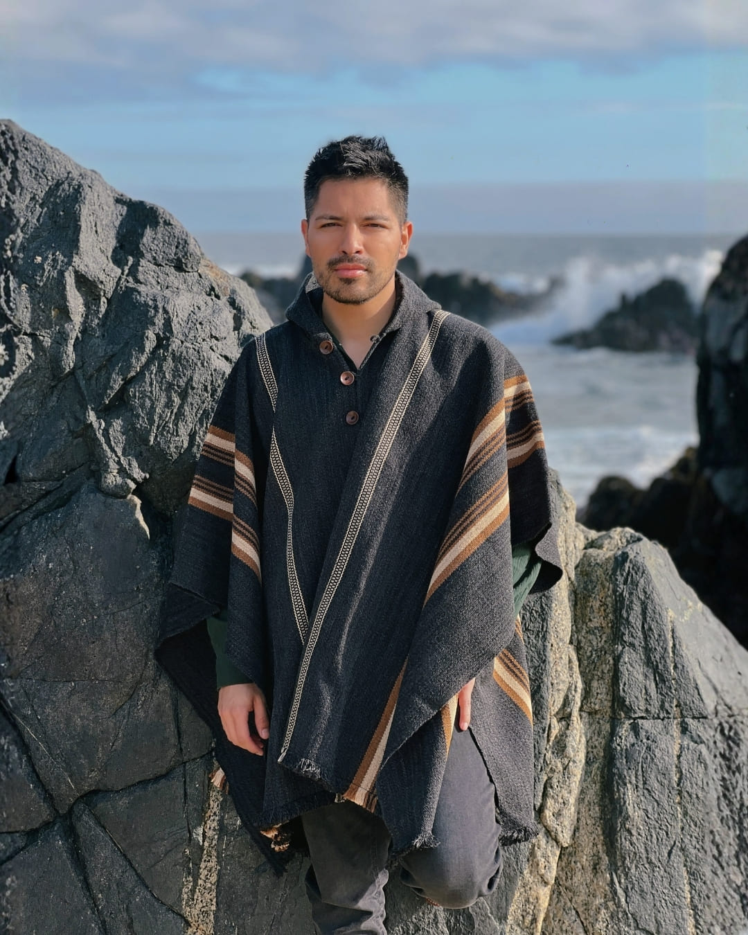 Hombre usando Poncho Arrayán gris marengo con líneas beige y camel en paisaje costero.